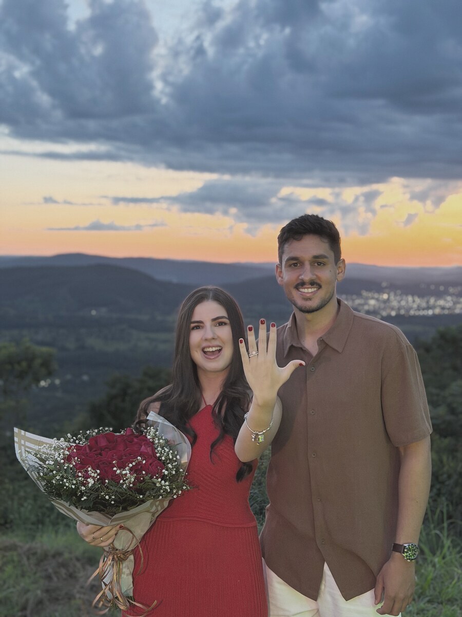 João e Mel ao pôr do sol num mirante — Mel mostra o anel de noivado segurando um buquê de rosas vermelhas.